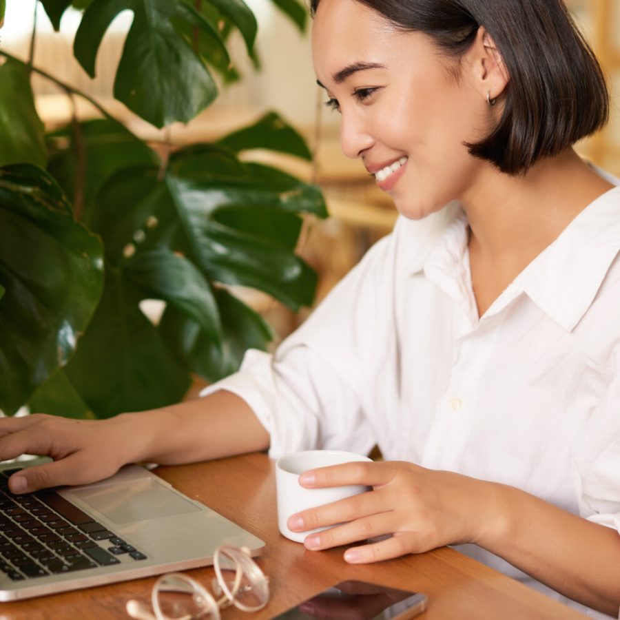 Student sitting in cafe with cup of coffee. Young asian woman working on laptop in restaurant, sitting with computer and smartphone.