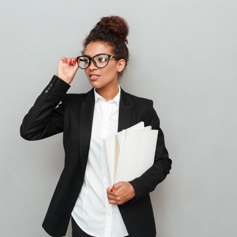 Image of young african business woman standing over grey wall looking aside holding documents.